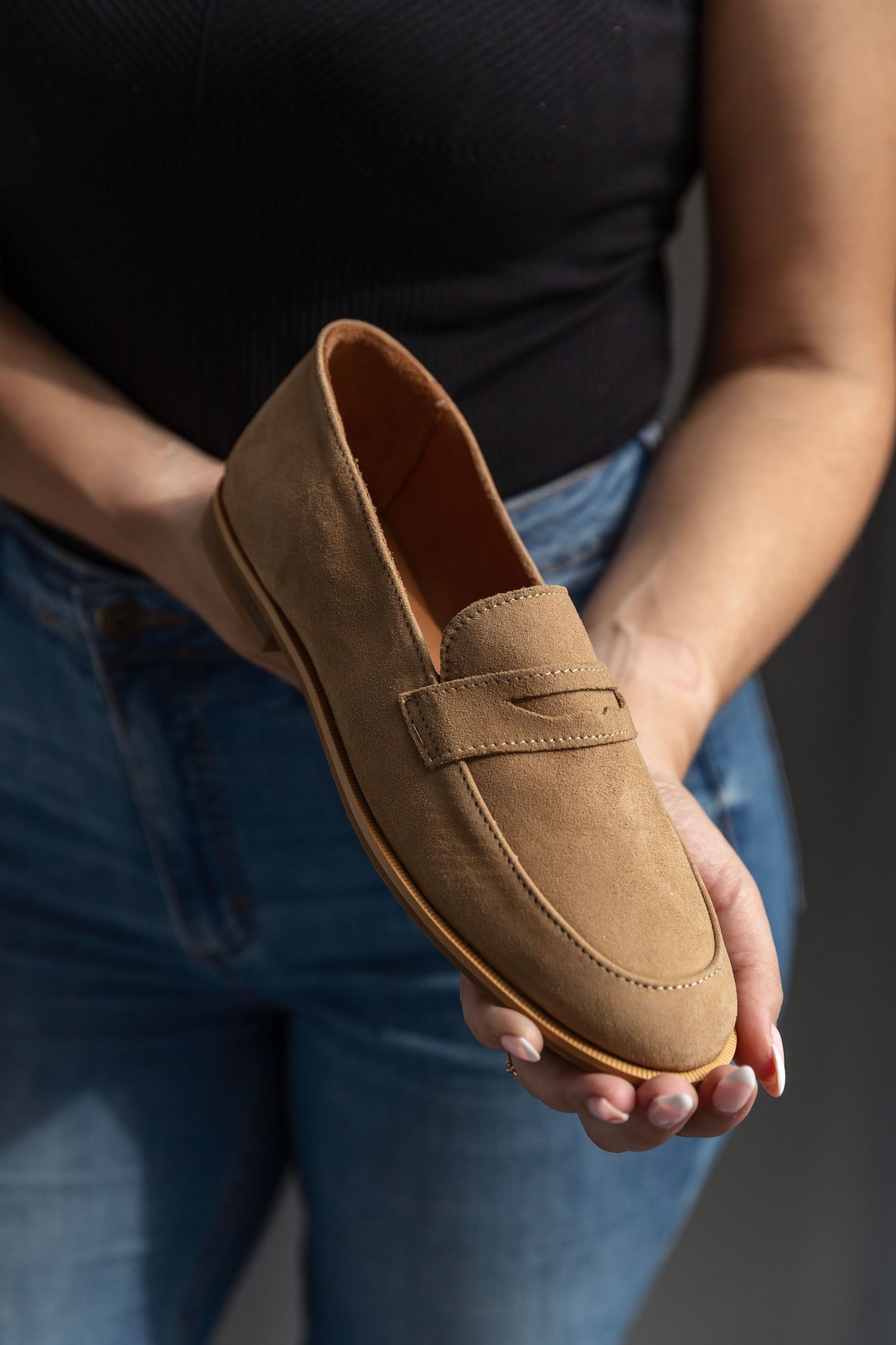 Woman holding a tan suede penny loafer with leather insole and rubber sole against grey background.
