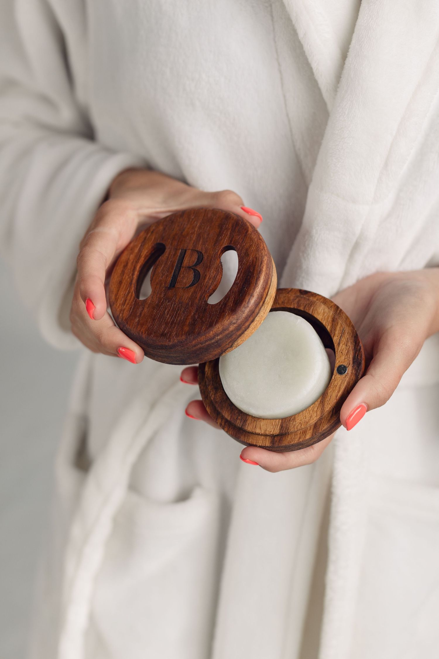 Hands with coral-colored nails hold an open wooden ring box with white interior against a white fabric background.