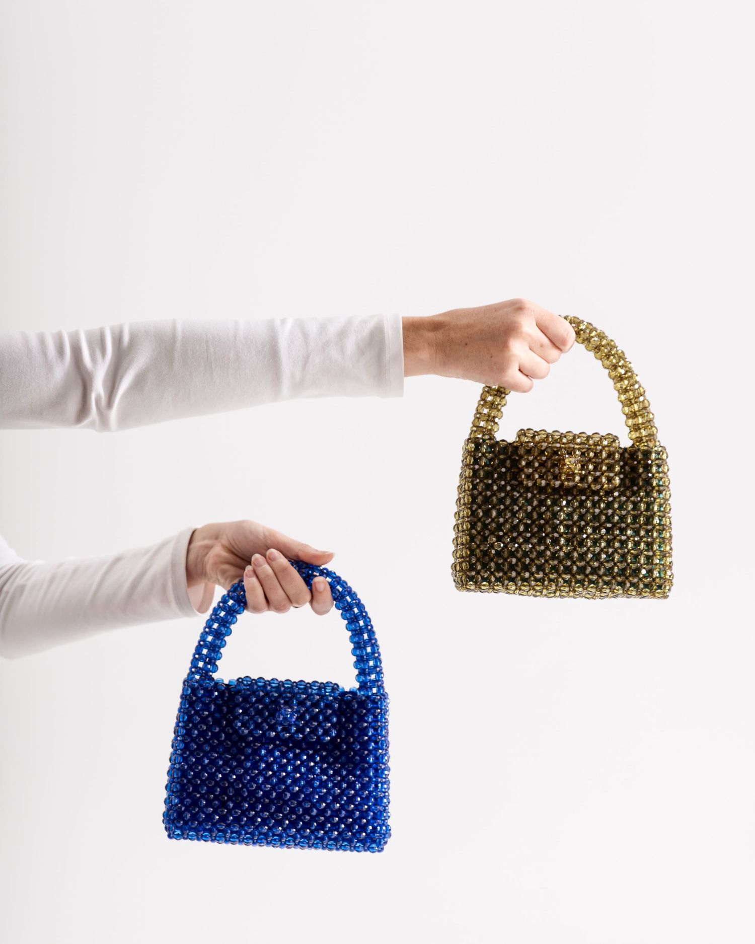 Two hands holding beaded handbags, one royal blue and one gold, against a white background.