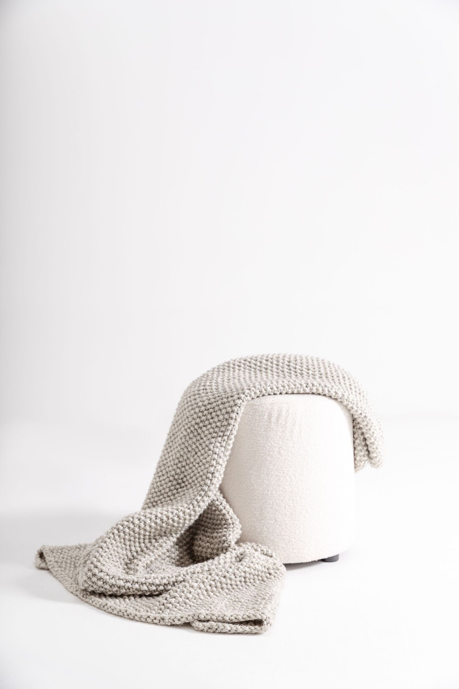 A cozy gray knitted throw blanket draped over a round white ottoman on a white background.