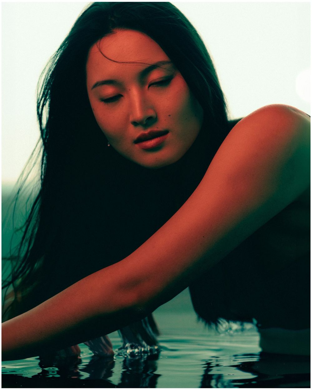 A young Asian woman with long dark hair leans gracefully over still water, eyes closed, in a serene poolside portrait.