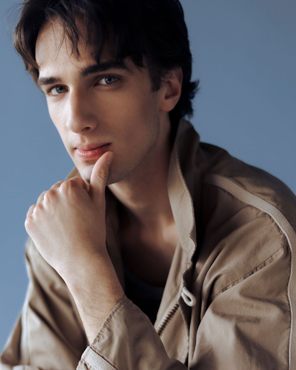 Young man with dark hair in beige jacket poses thoughtfully with hand on chin against blue background.