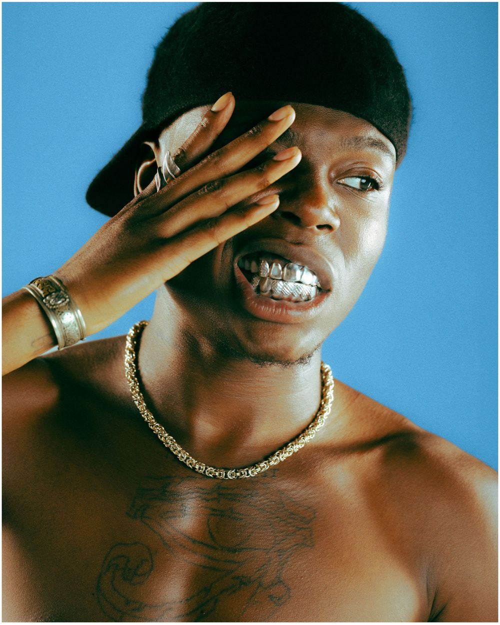 Shirtless man with gold grillz, chain necklace, bracelet, and black cap covering one eye against blue background.