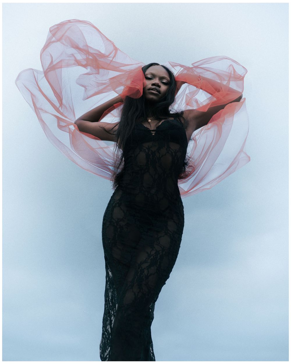A Black woman in a black lace gown poses dramatically with flowing red fabric against a light gray background.