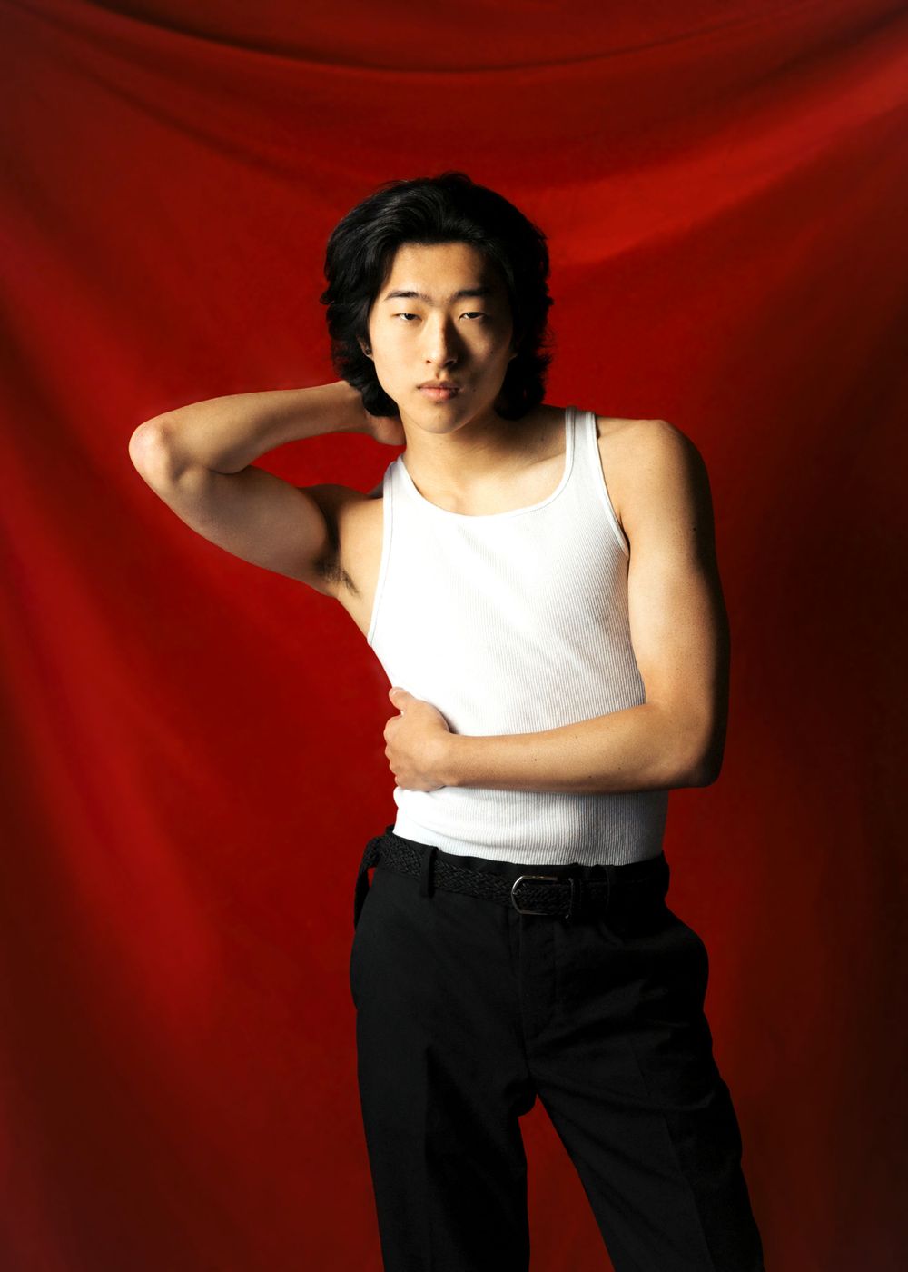 Person in a white tank top and dark pants poses against a red backdrop with one hand behind their neck.