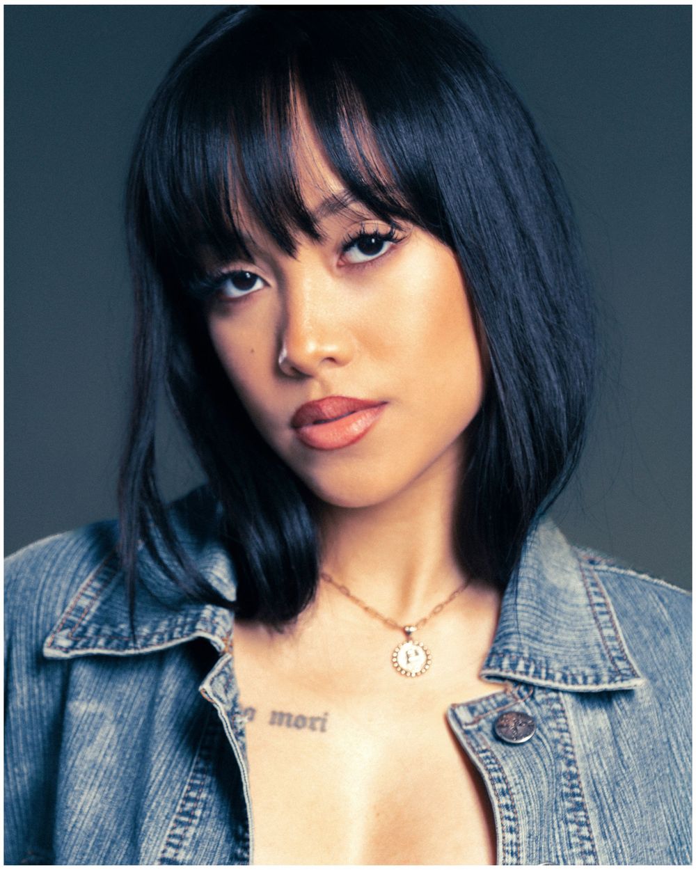 Portrait of someone with black layered hair and bangs wearing a denim jacket against a dark backdrop.
