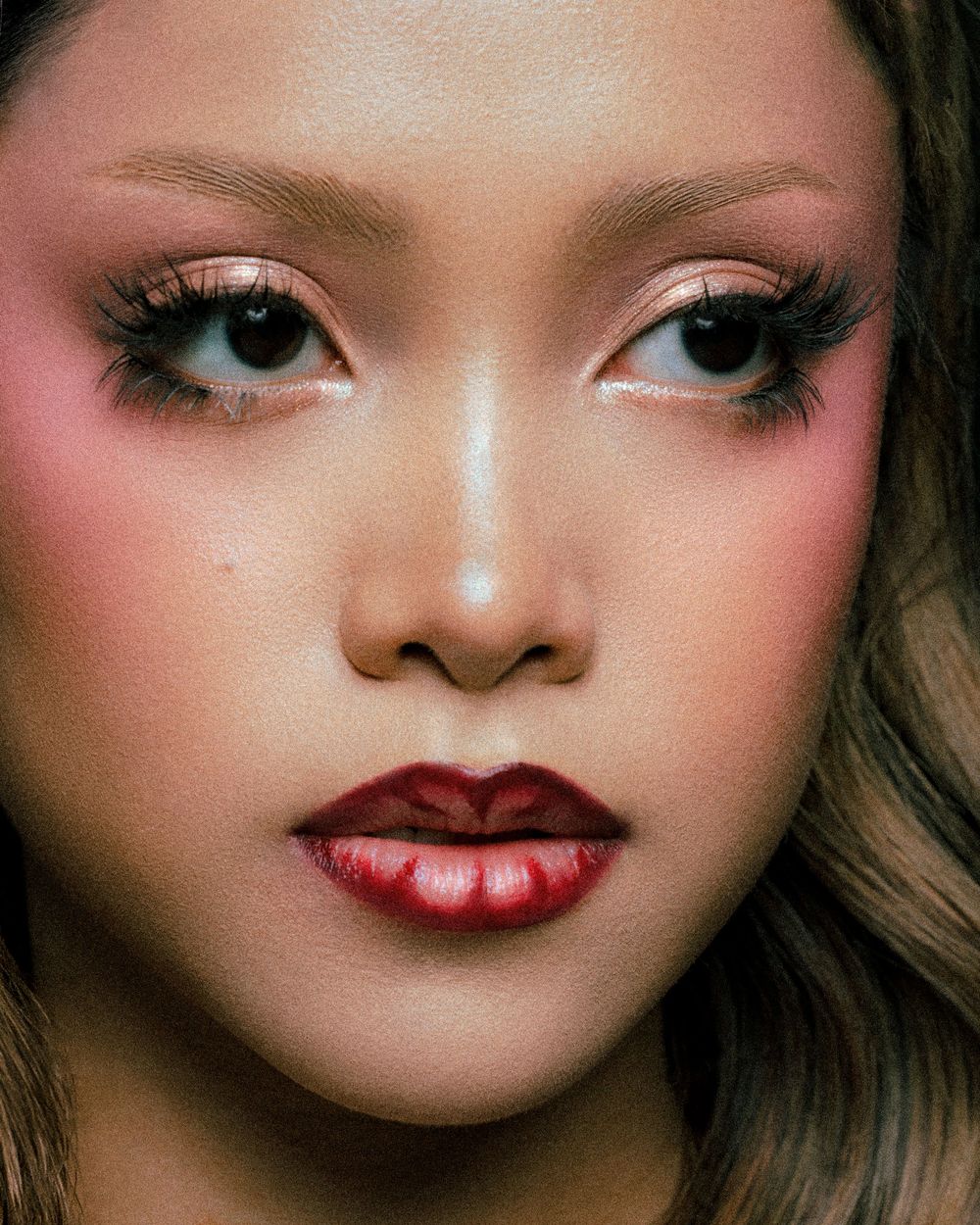 Close-up portrait of a woman wearing bold red lips, rosy blush, and dramatic lashes with shimmer eyeshadow.