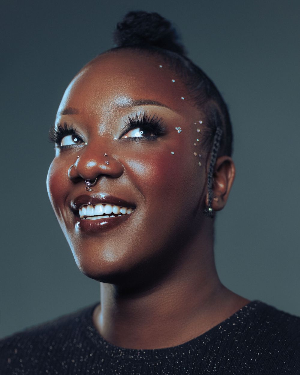 Smiling young woman with glitter face gems, nose ring, braids, and a bun hairstyle against a dark background.