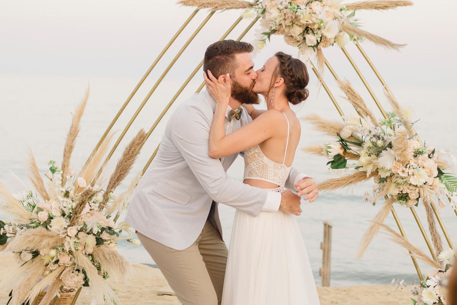 A romantic beach wedding ceremony under a bamboo arch decorated with pampas grass and white floral arrangements.