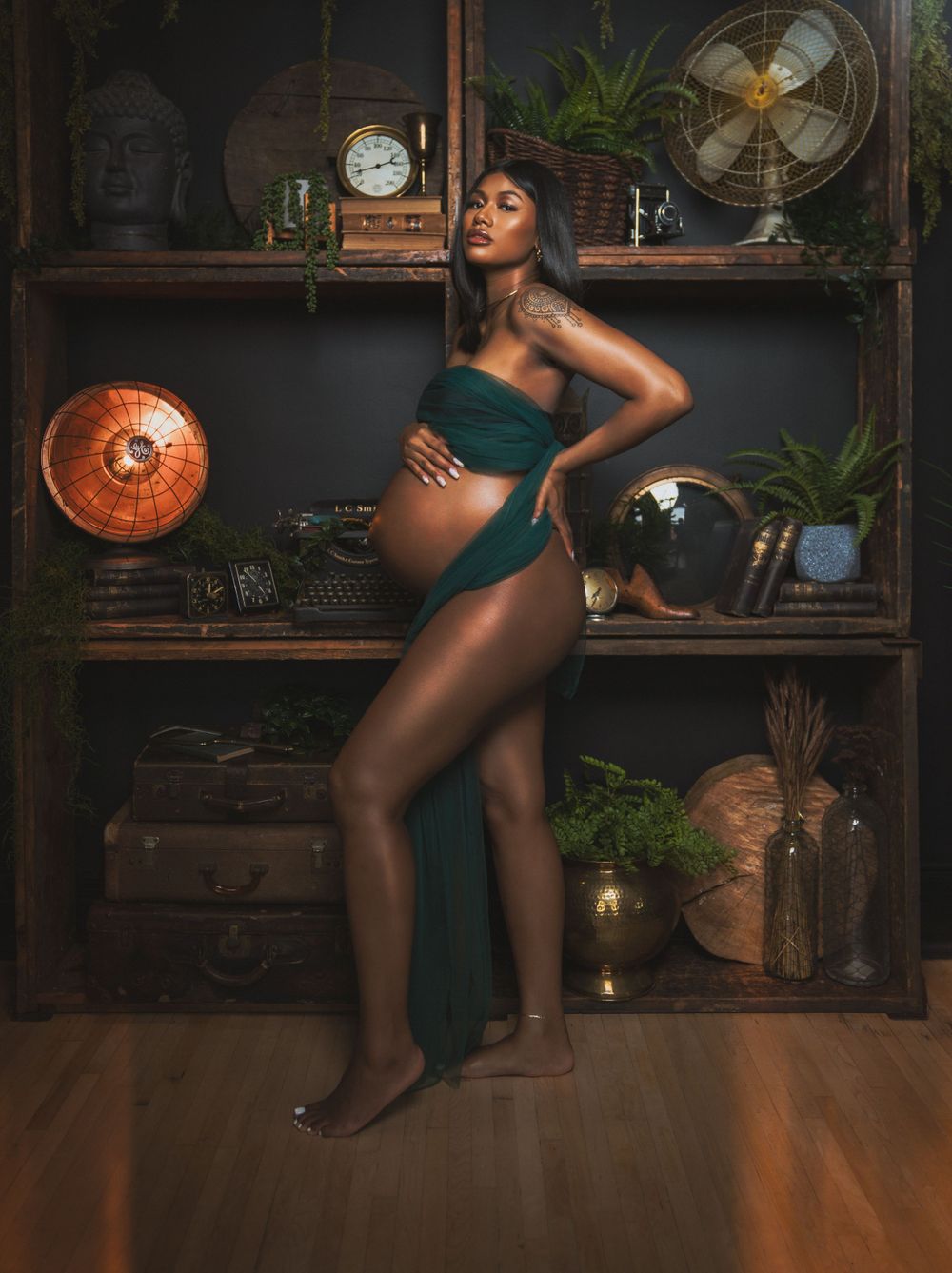 Pregnant Black woman in emerald green wrap posing before vintage styled bookshelf backdrop for her fashion maternity session in Cincinnati