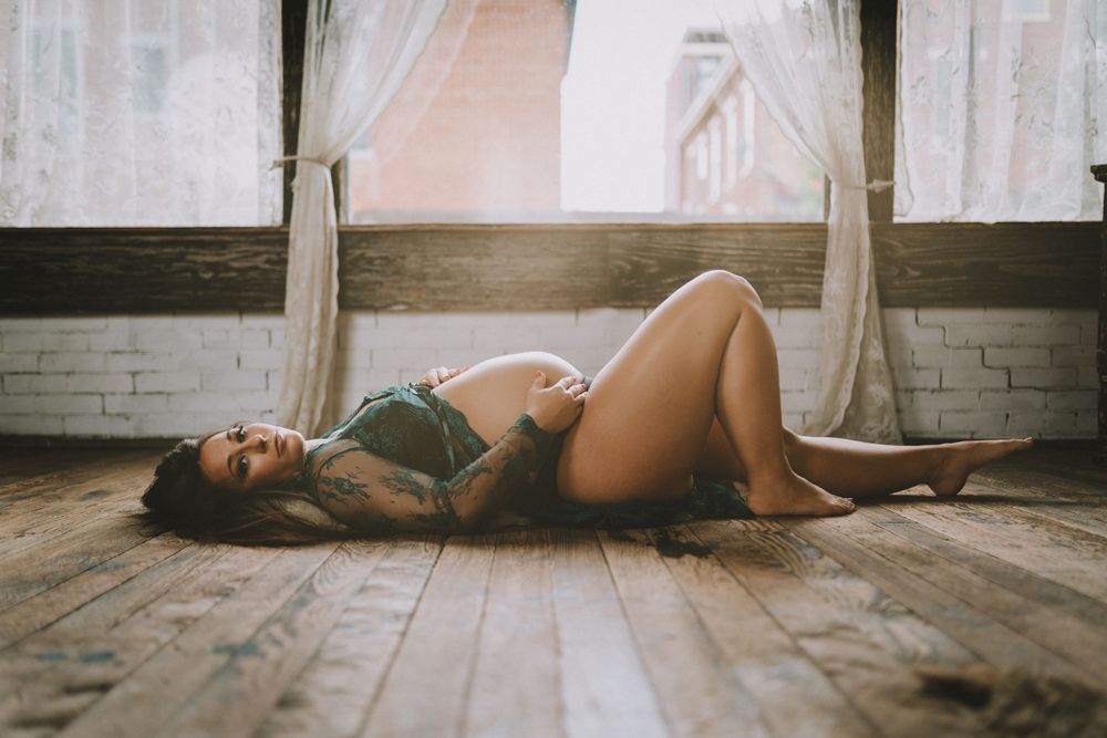 Pregnant woman in green lingerie lying on hardwood floor near sunlit windows in artistic maternity photo.