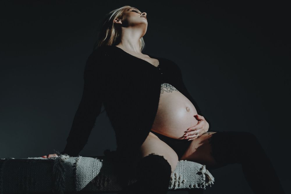 Pregnant blonde woman in black bodysuit sitting in dramatic dark low-key lighting for her maternity session in Cincinnati
