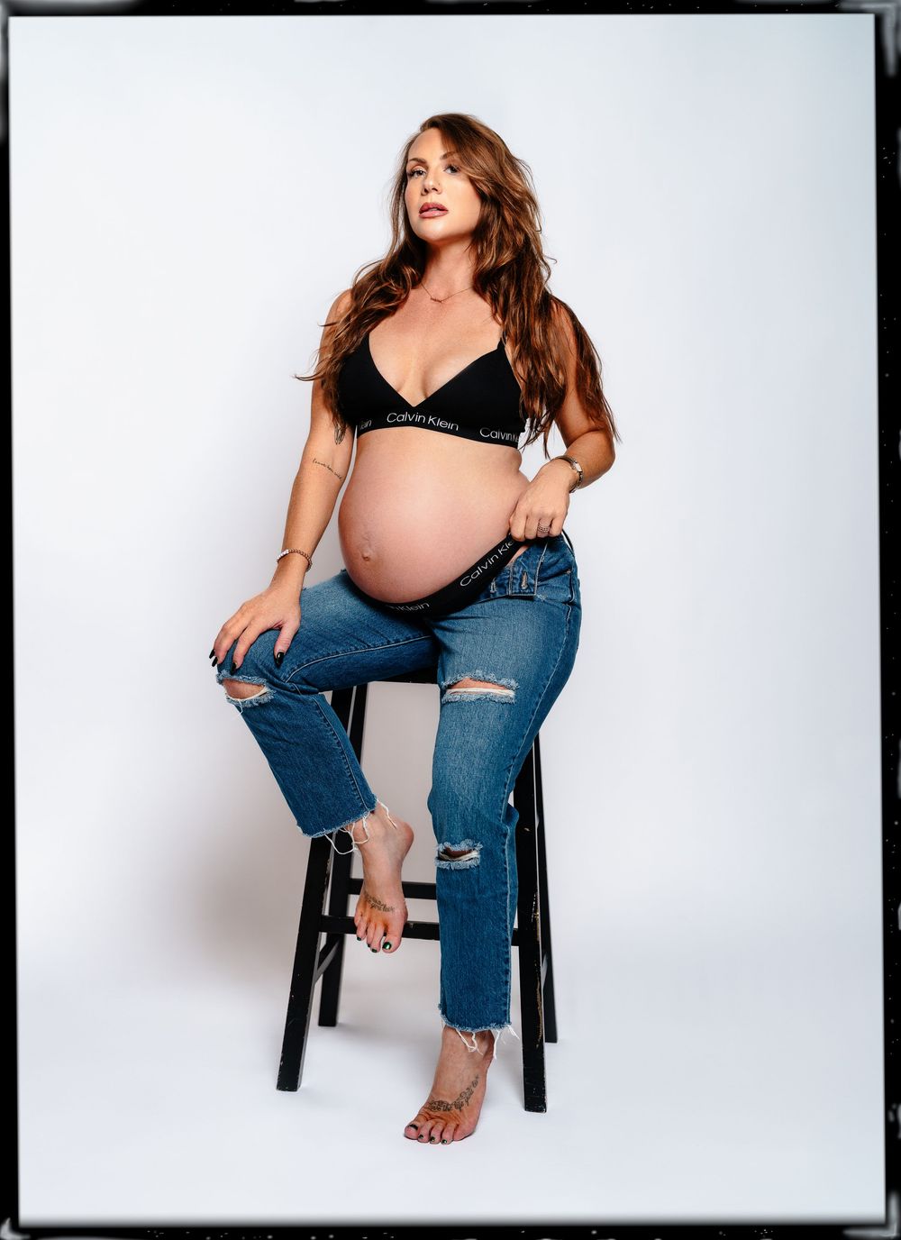 Red haired pregnant woman posing in jeans and bra sitting on a black stool in a studio maternity session in Cincinnati Ohio