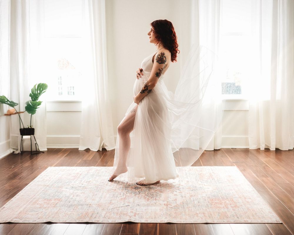 A tattooed pregnant woman in a flowing white sheer gown stands gracefully by sunlit windows indoors in Cincinnati