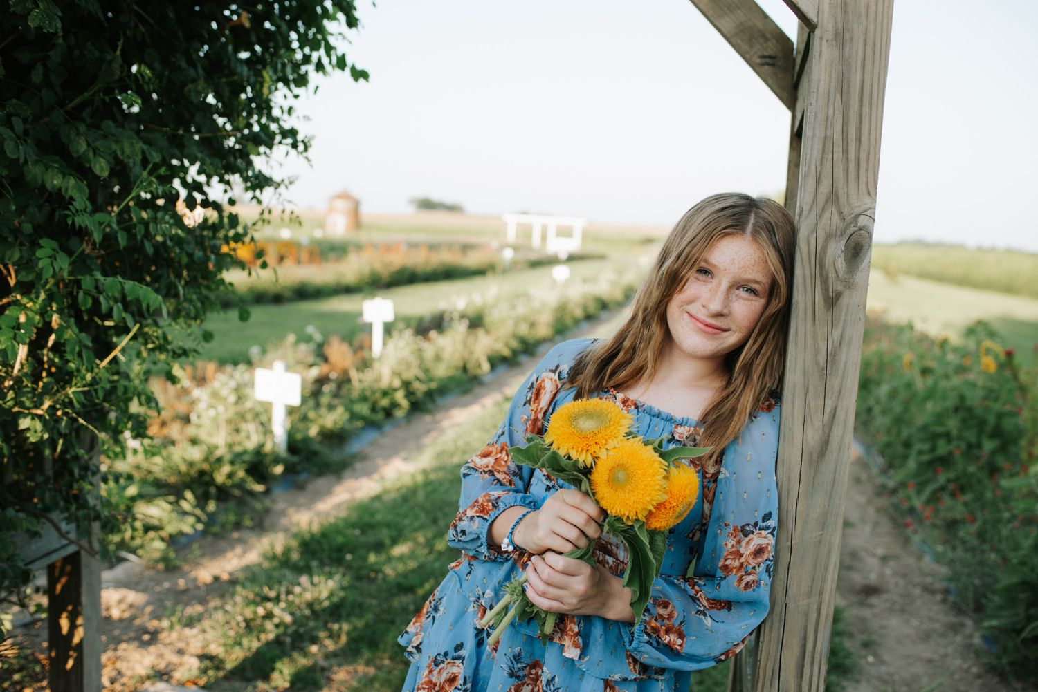 A person in a blue floral dress holds yellow autumn leaves while posing against a rustic wooden post outdoors at Flower Farm photoshoot.