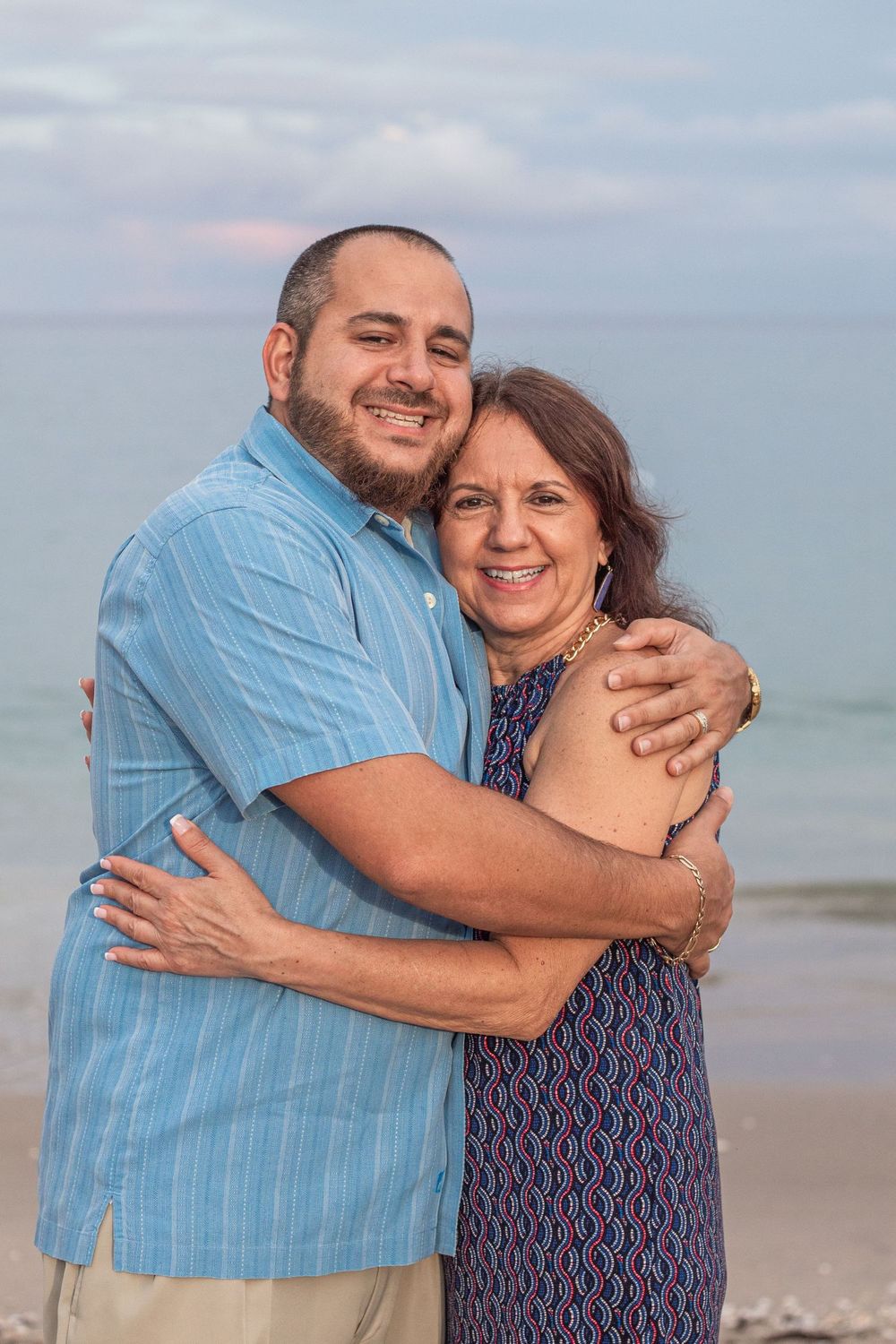 A man and older woman embrace and smile on a beach at dusk, wearing casual summer attire at Lake Worth Beach.