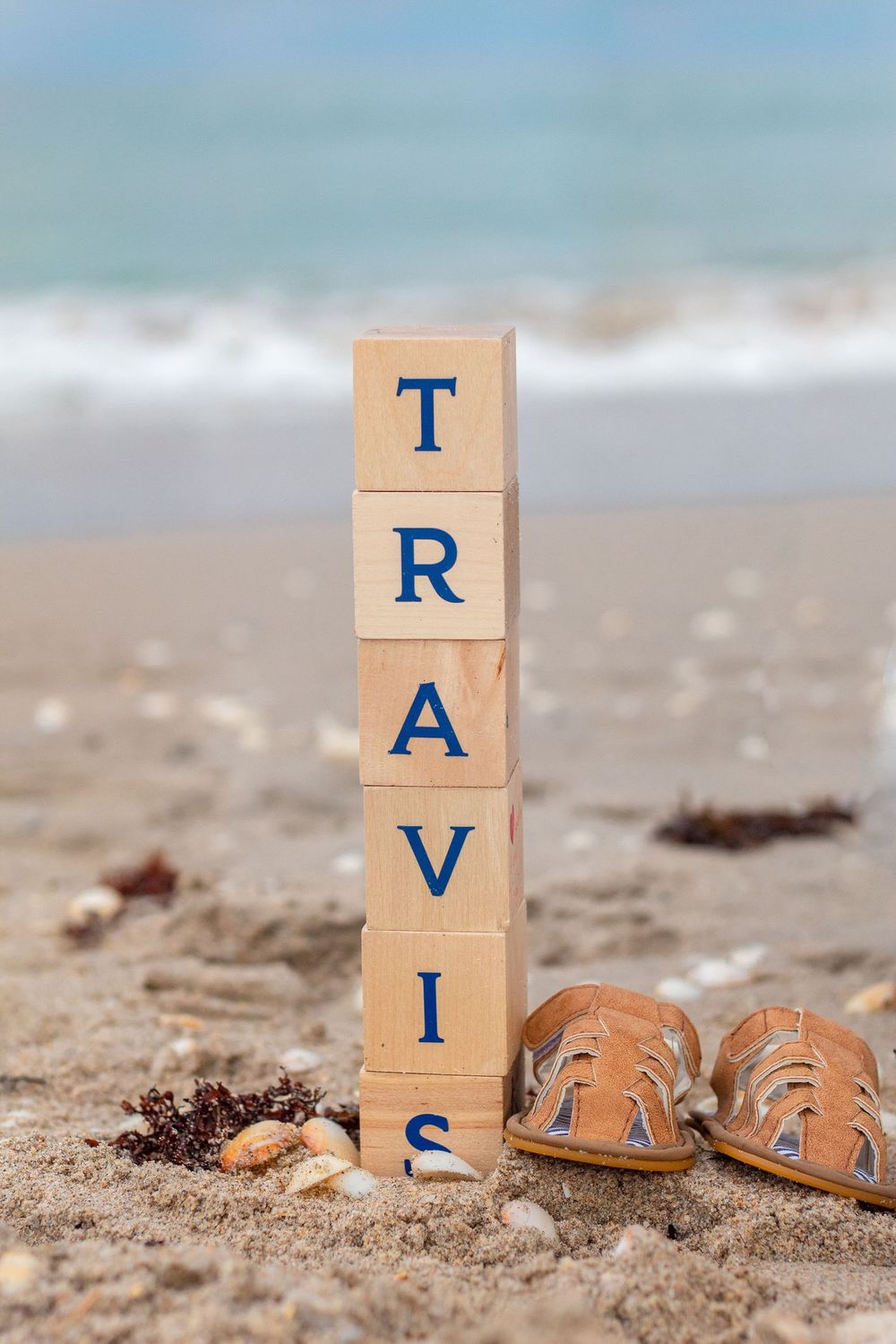 Wooden alphabet blocks spelling TRAVIS stacked on sandy beach with sandals and seashells, ocean waves in background at Lake Worth Beach.