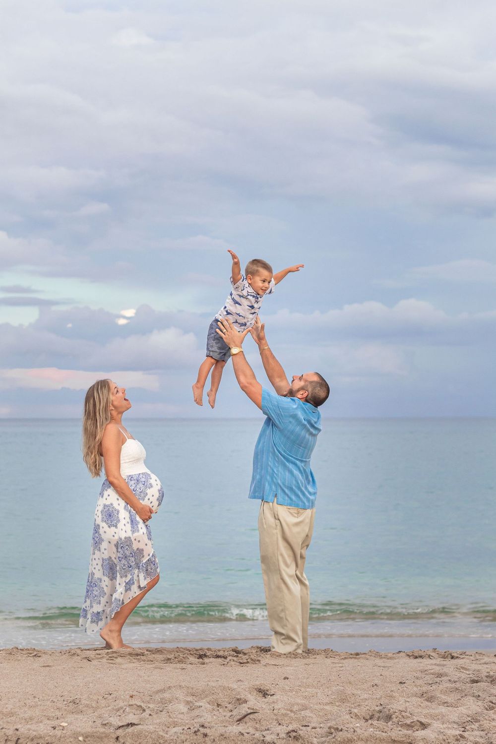 Pregnant woman and man tossing toddler in the air on a beach at dusk, family maternity photo session at Lake Worth Beach.
