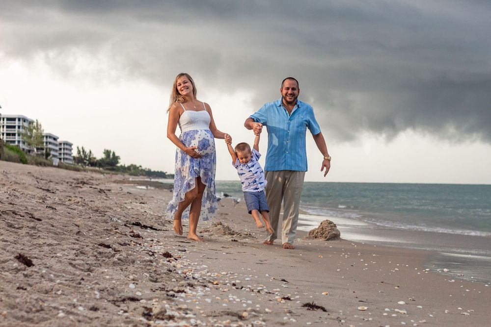 Family of three walking on beach, parents swinging child, stormy sky backdrop, ocean waves, sandy shore at Lake Worth Beach.
