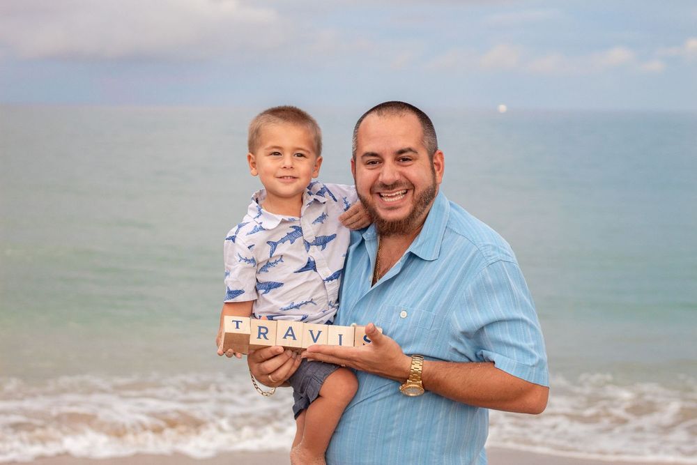 A man and young boy smile together on a beach holding Scrabble tiles spelling TRAVIS with ocean waves behind them at Lake Worth Beach.