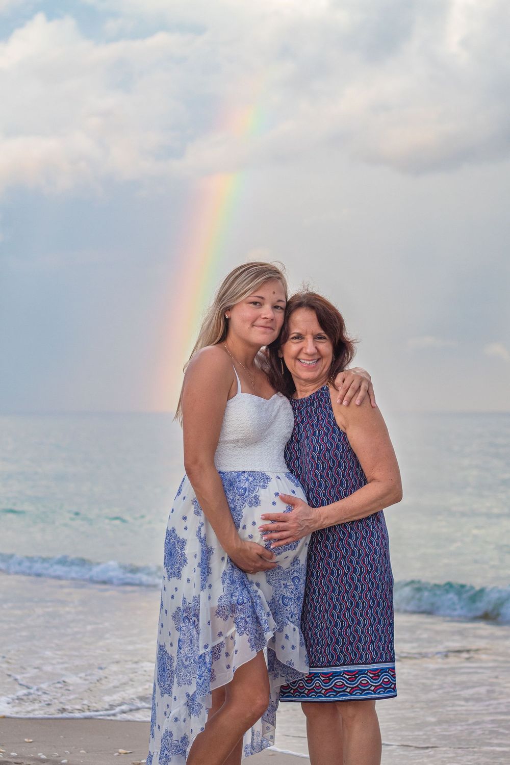 Pregnant woman and older woman embracing on beach with rainbow over ocean behind them at Lake Worth Beach.