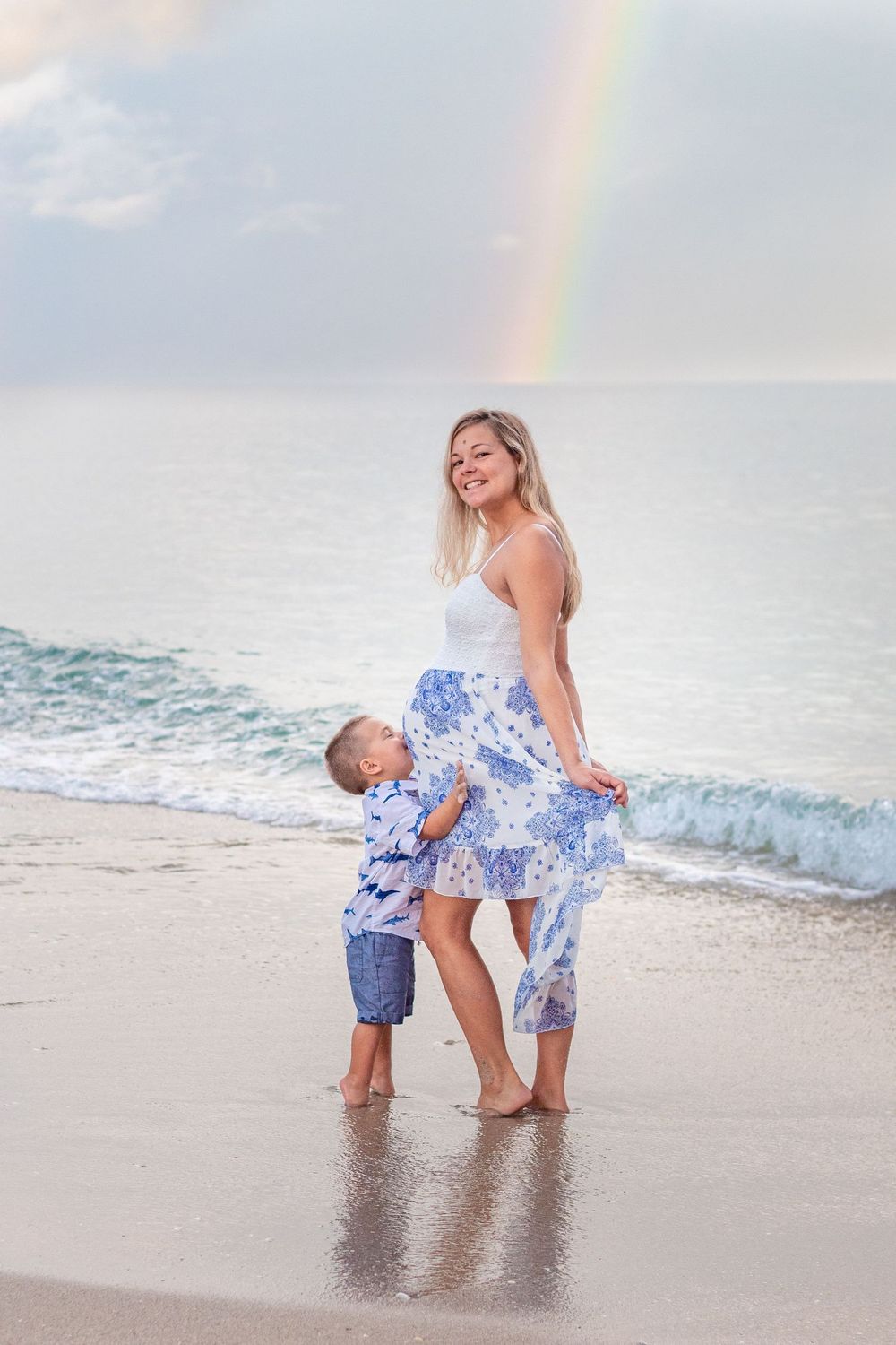 A smiling woman and young boy stand on a beach shoreline with a rainbow over the ocean behind them at Lake Worth Beach.