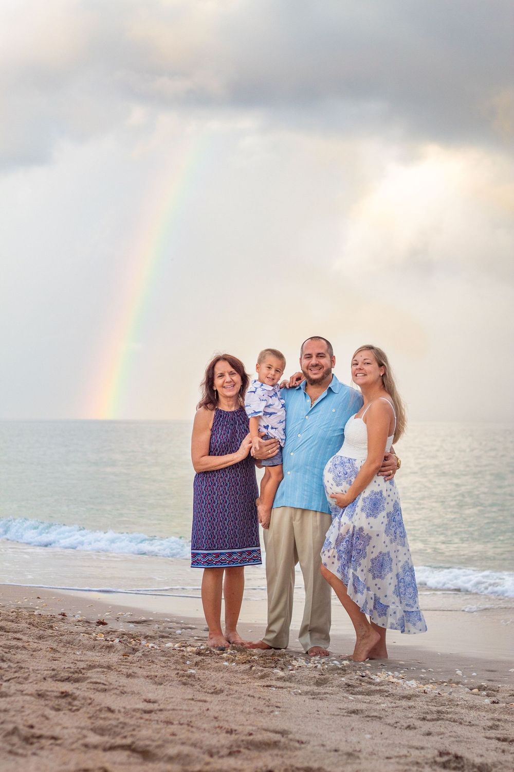 Family of four poses on a sandy beach with a vibrant rainbow over the ocean in the background at Lake Worth Beach.