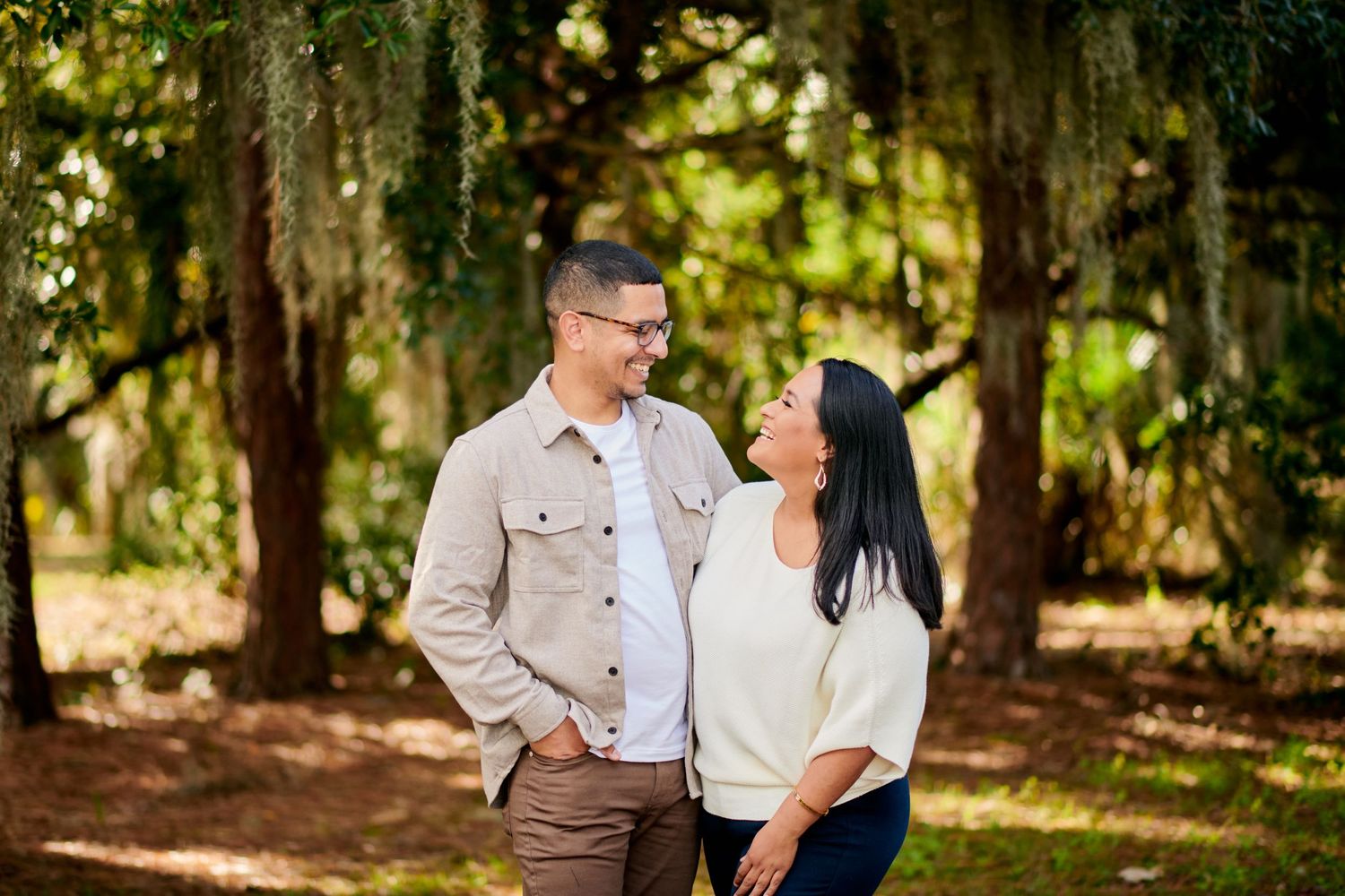 A couple shares a tender moment while standing together in a sunlit wooded area with Spanish moss in the background.