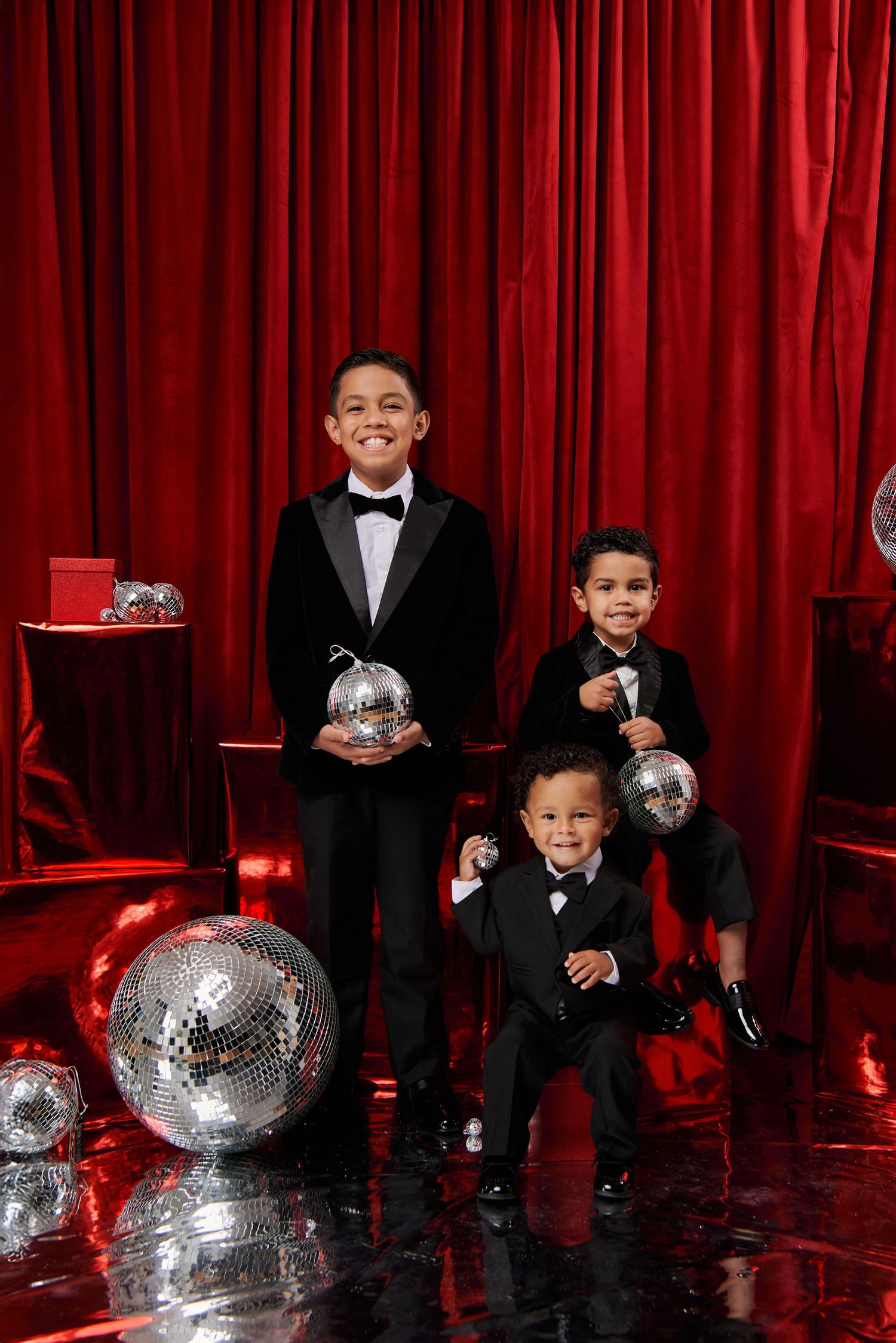 Three children in formal black tuxedos pose with silver disco balls against a dramatic red curtain backdrop.