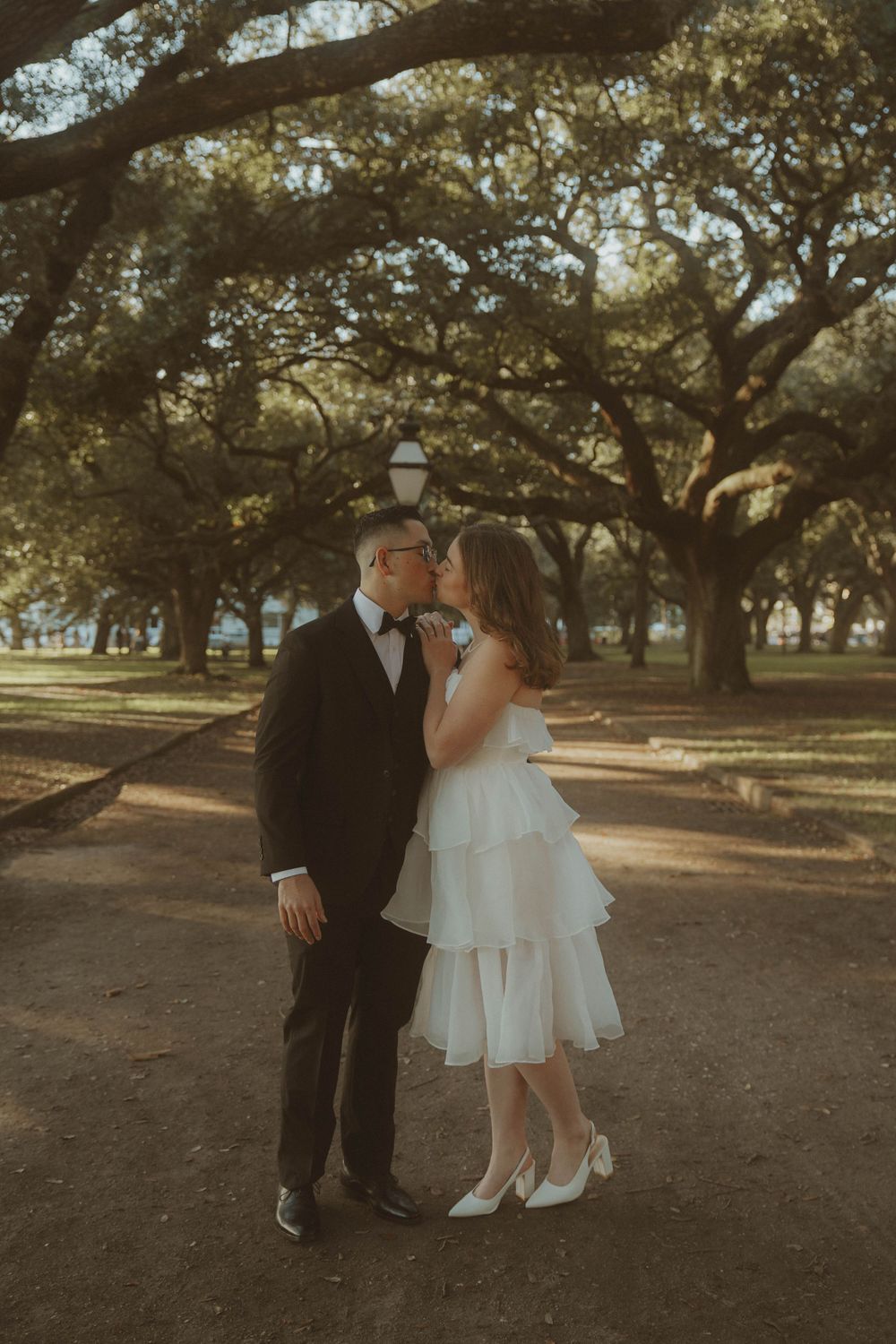 A romantic wedding couple poses under majestic oak trees in a park during golden hour.