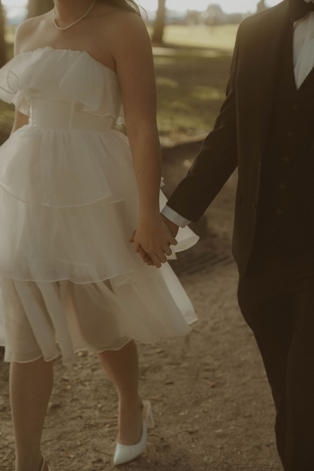 A close-up view of a couple holding hands, showing her tiered white dress and his black suit.
