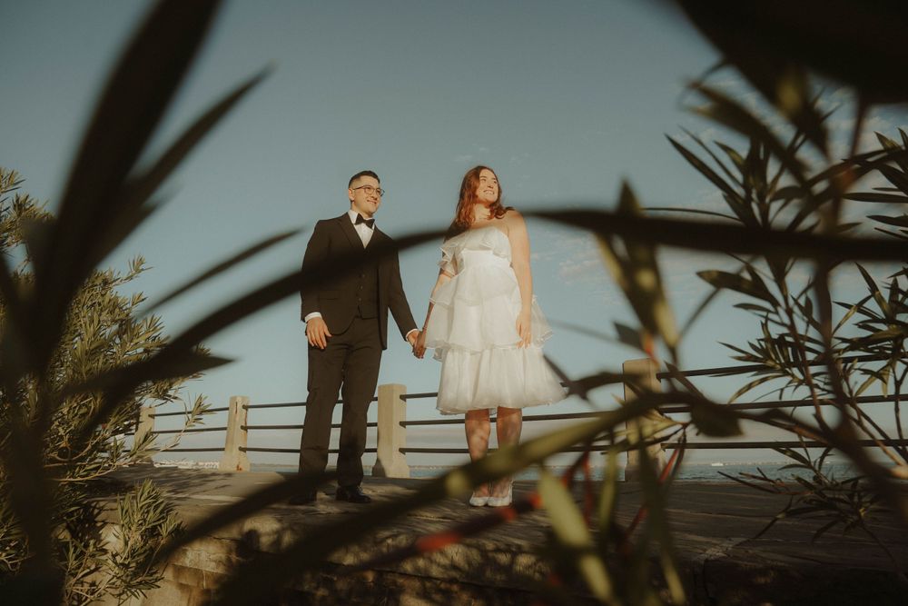 A couple holding hands while standing on a coastal path with plants in the foreground during sunset.