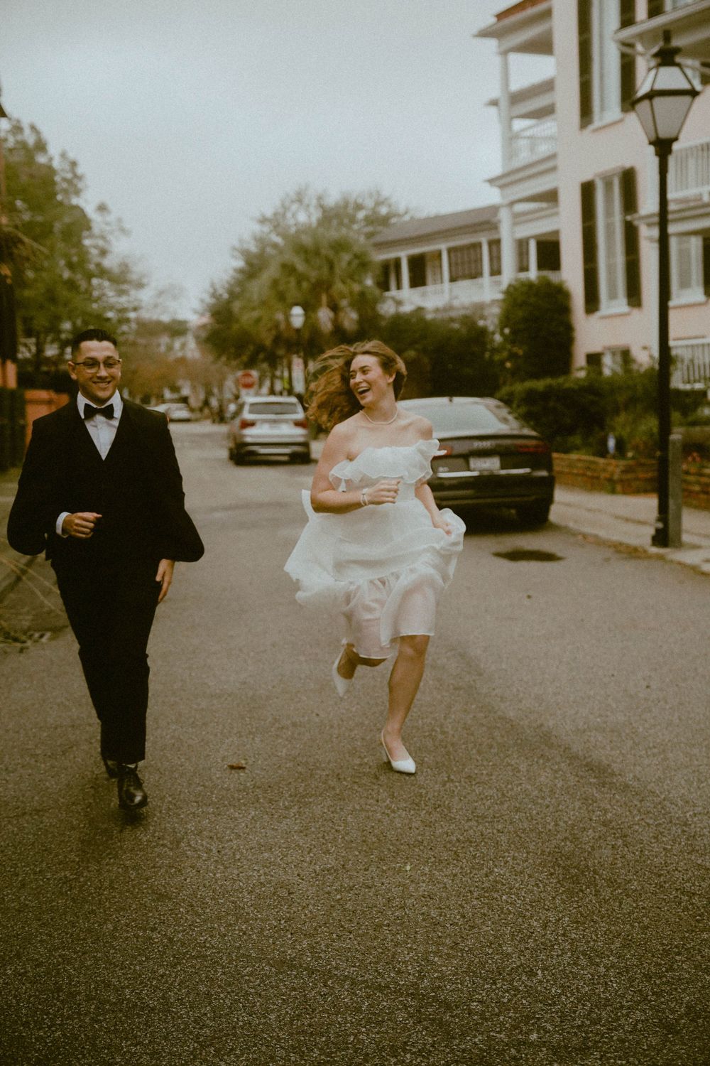 A couple in wedding attire runs playfully down a historic Charleston street with classic architecture and parked cars.