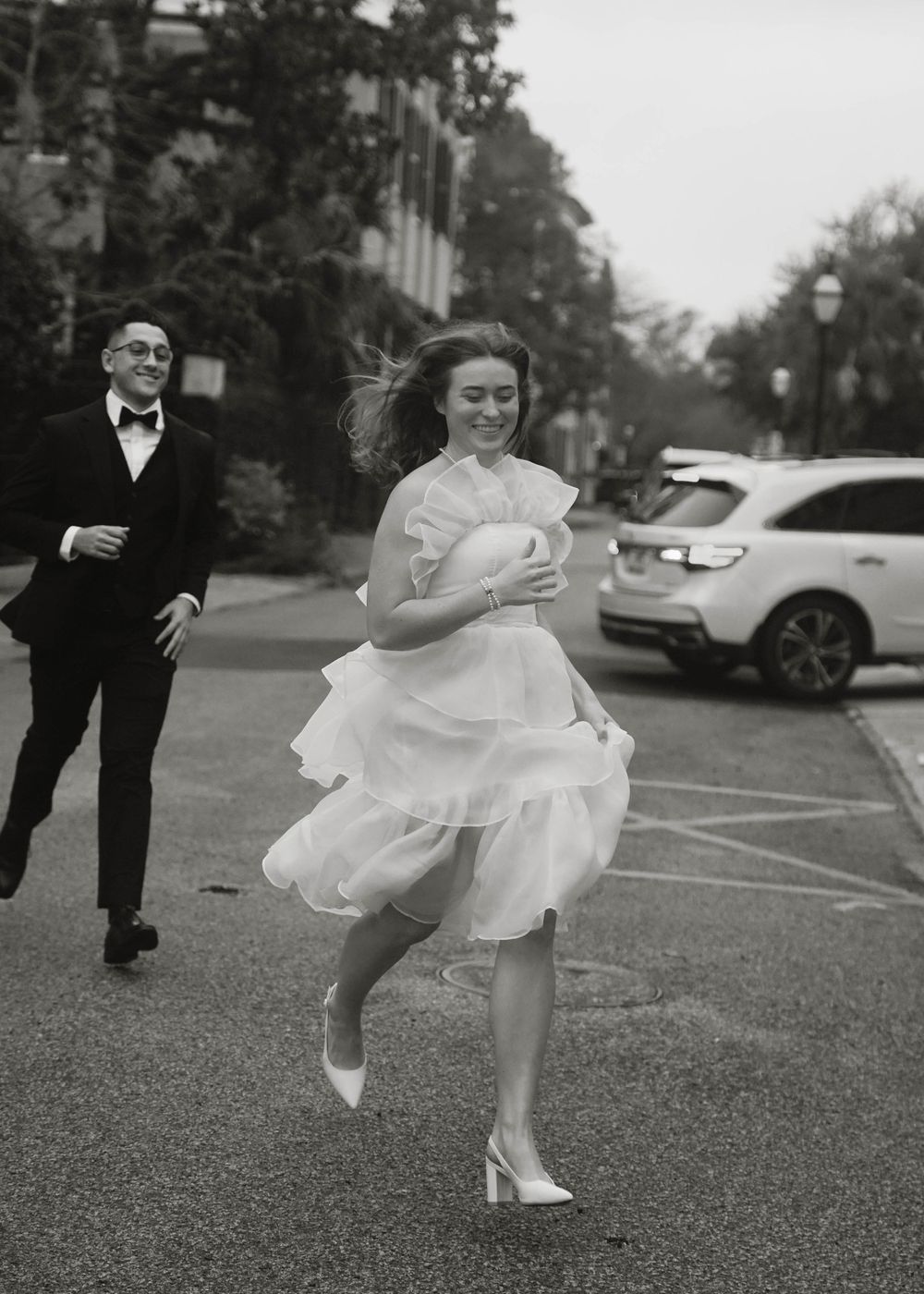 A black and white photo of a couple in formal attire running through a parking lot with flowing dress movement.