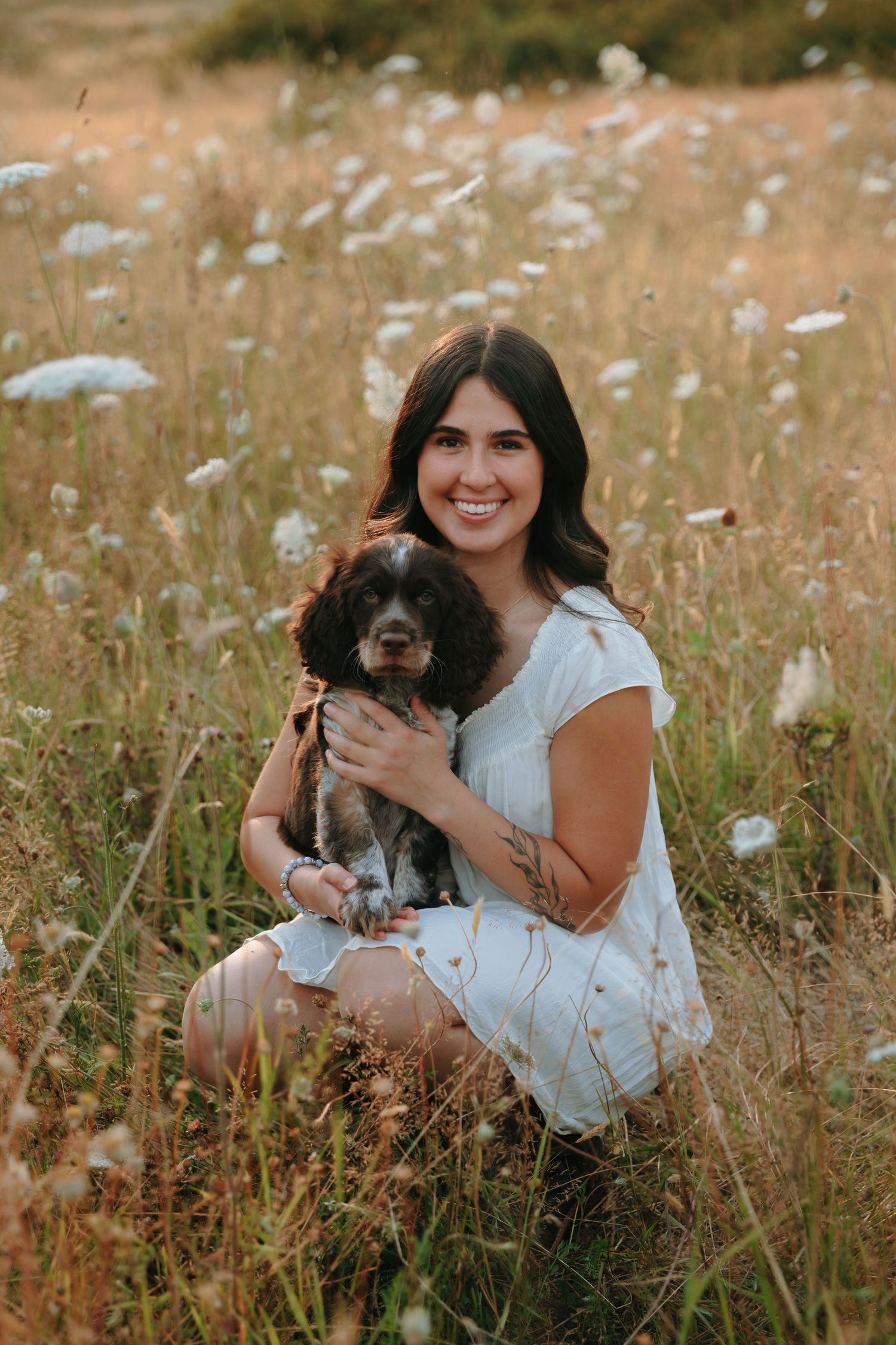 Woman in white dress sitting in golden field of wildflowers while holding small brown curly-haired dog.