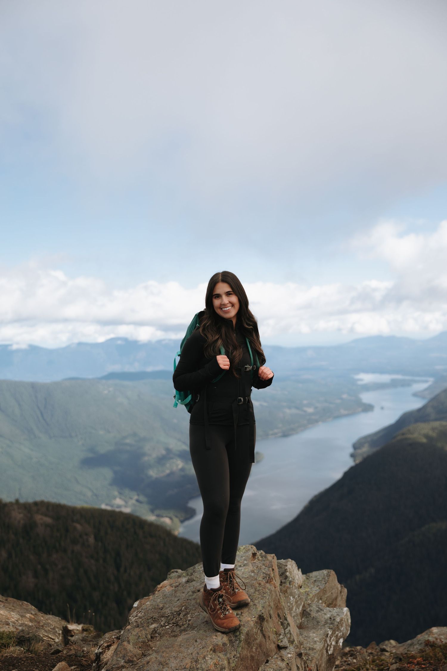A solo hiker stands on a mountain summit overlooking a scenic lake and mountain vista in the distance.