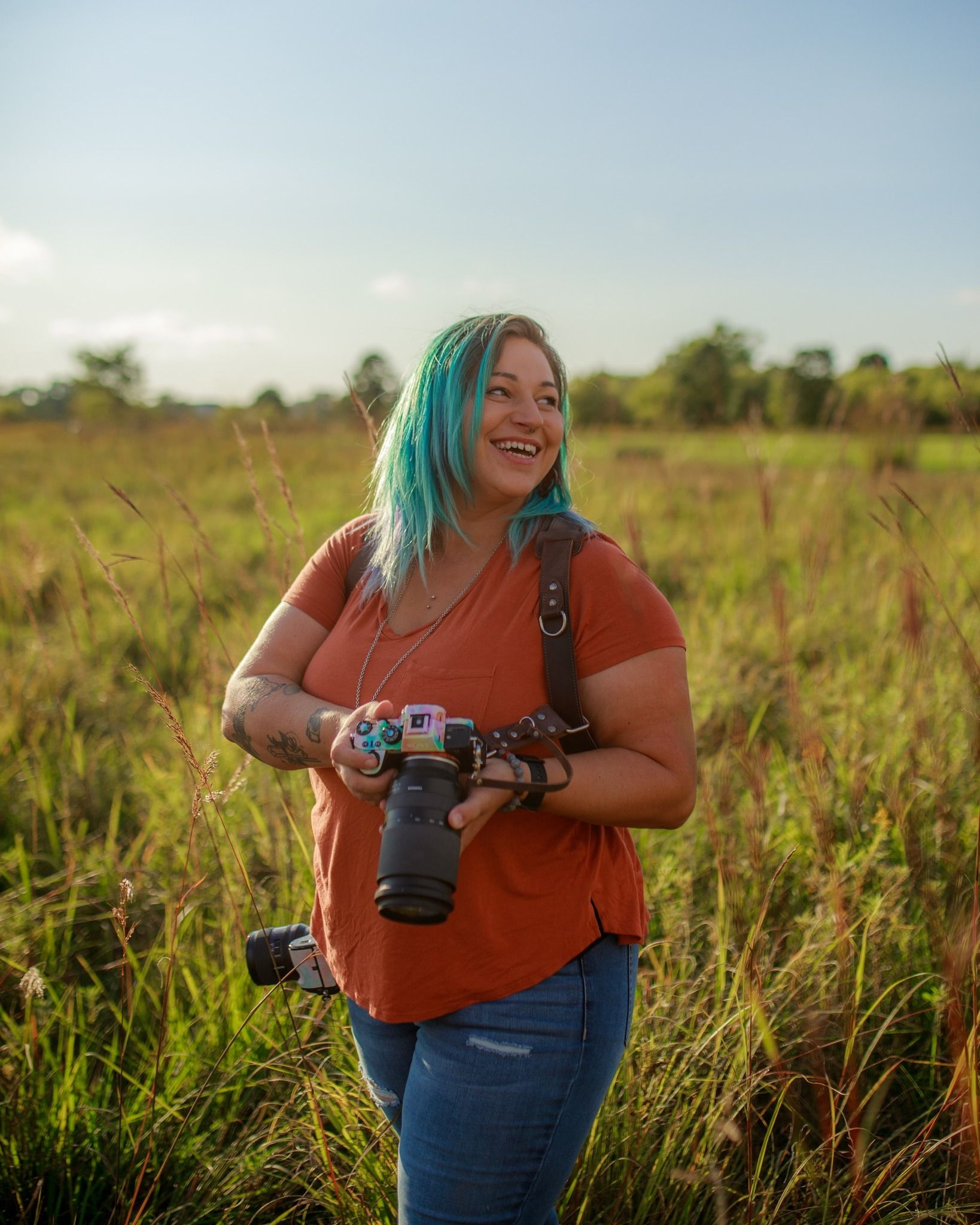 Photographer in orange shirt holds camera while standing in sunny meadow during golden hour.
