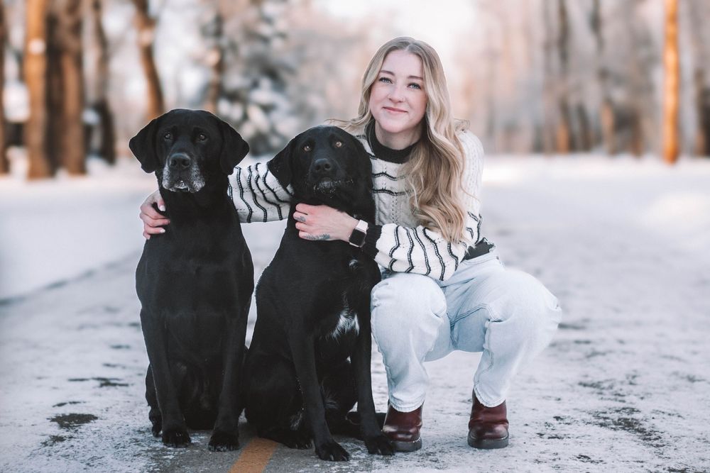 Smiling blonde woman crouching between two black Labrador Retrievers on a snow-covered park path in winter.
