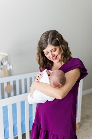 In a nursery with a blue crib, a person in a purple dress lovingly holds a swaddled newborn baby against their chest.
