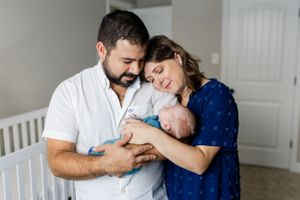 Parents in white and blue outfits tenderly care for their newborn baby near a white crib in a nursery setting.