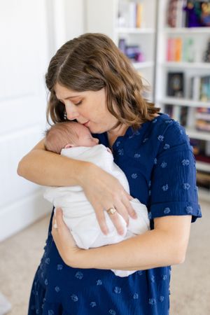 Loving parent in navy blue dress tenderly holding and embracing newborn baby wrapped in white blanket by window.