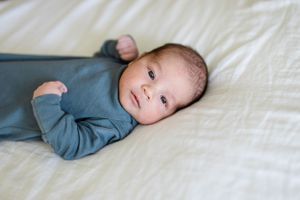 A baby wearing a soft blue sleeper rests on white bedding while gazing at the camera.