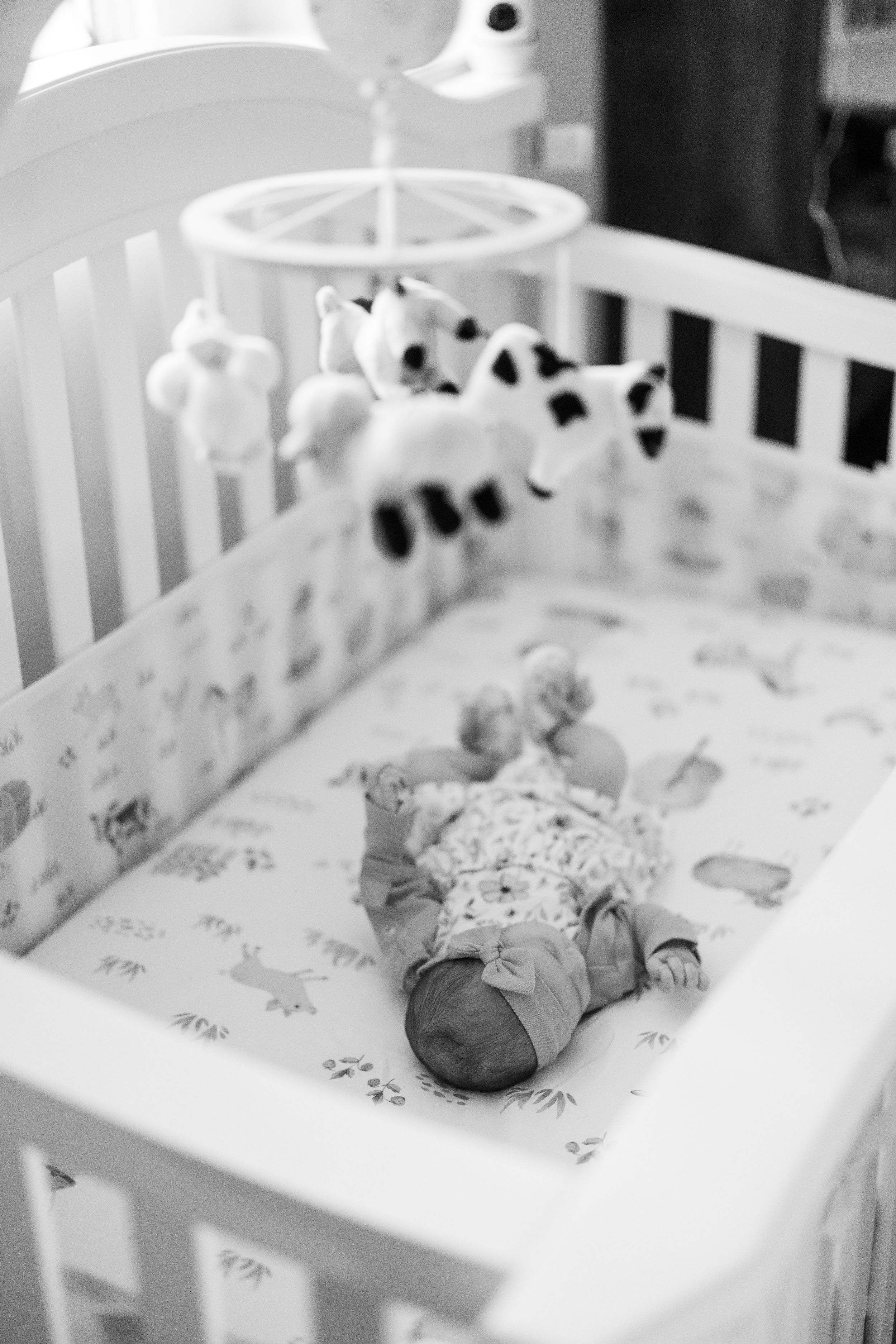 A black and white photo of a baby lying in a crib with a spotted mobile hanging above.