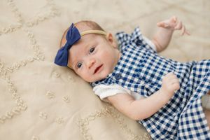 A baby in a blue gingham dress and matching hair bow lies on a cream-colored textured surface.