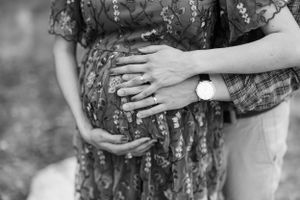 Black and white photo showing hands embracing a pregnant belly through a floral dress, with a wristwatch visible.