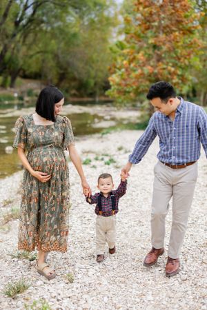 A family walks together on a gravel path near water, with autumn trees and foliage in the background.