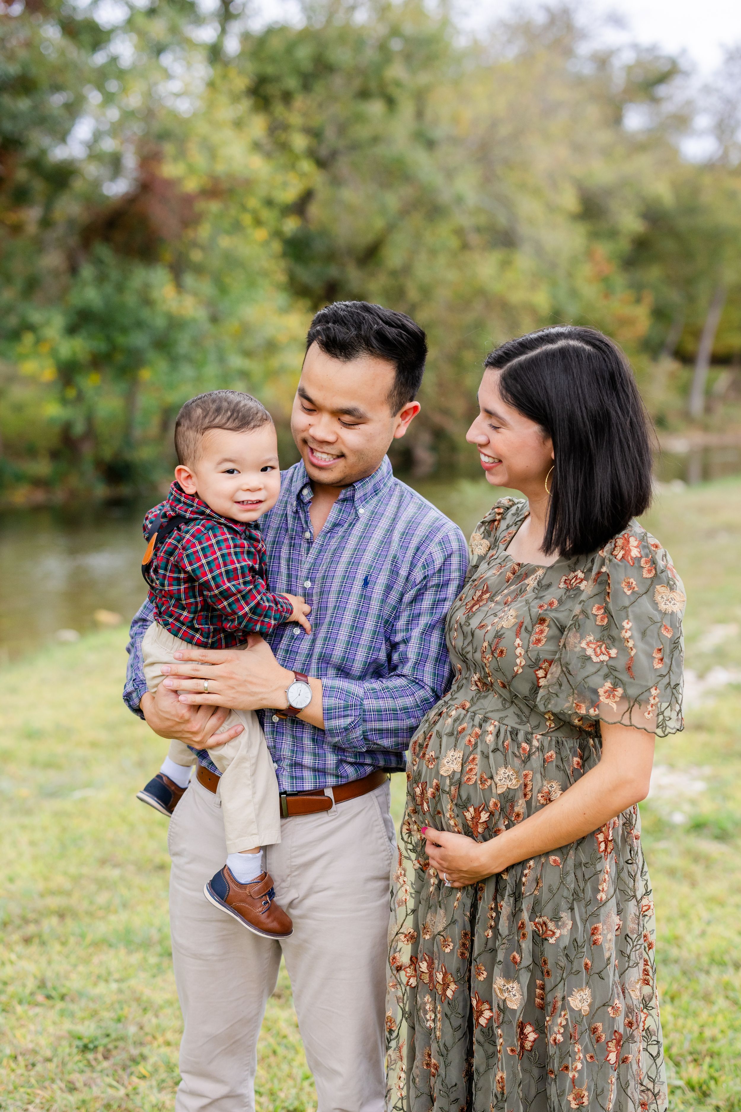 Expecting family shares a tender moment by a pond with their young child while enjoying the autumn scenery.