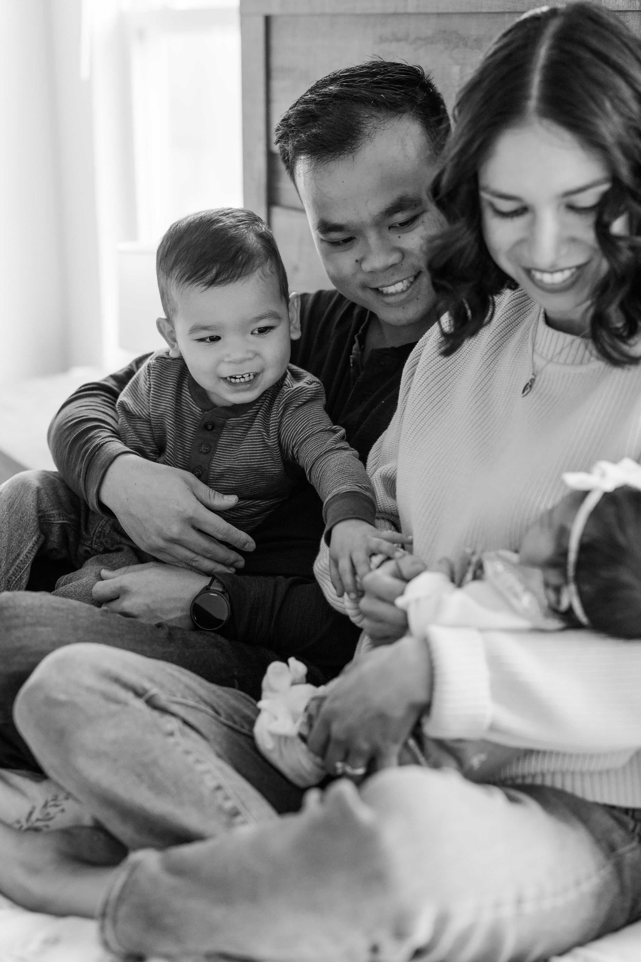 Black and white intimate family portrait showing parents and two small children sharing a tender moment together on a bed.