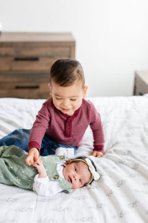 Two young children in casual clothing interact while laying on white bedding in a bright minimalist bedroom setting.