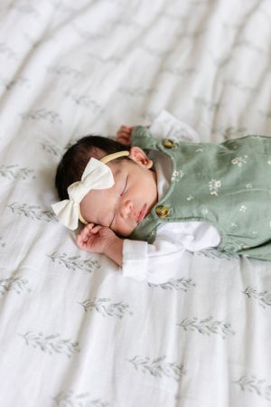 Sleeping newborn wearing sage green outfit and white bow headband rests on floral patterned bedding.